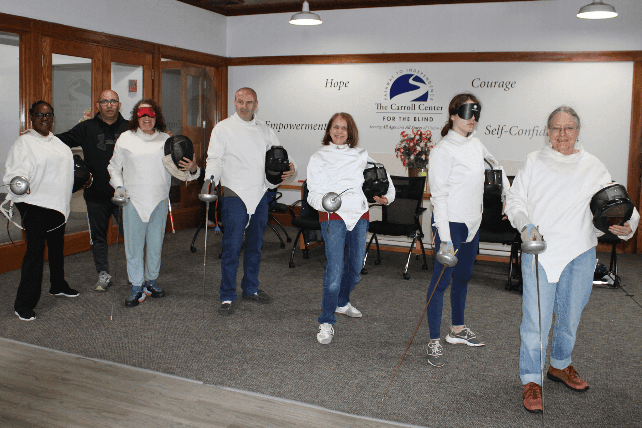 Six blind fencers and an instructor pose indoors at The Carroll Center for the Blind, holding foils in stance during a fencing activity.