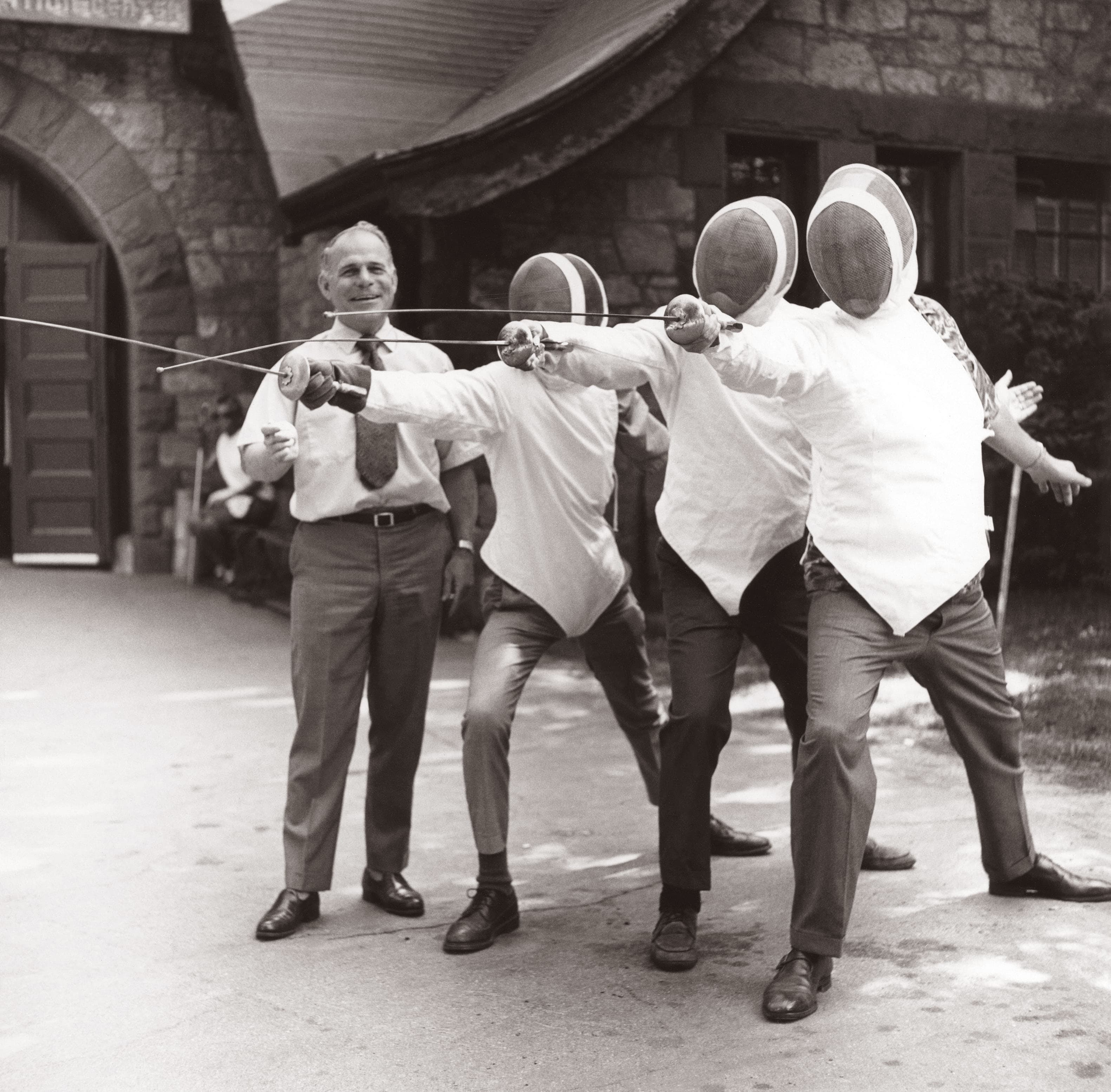 Three blind fencers in masks and gear practice outdoors with a smiling instructor guiding them in front of a historic stone building.