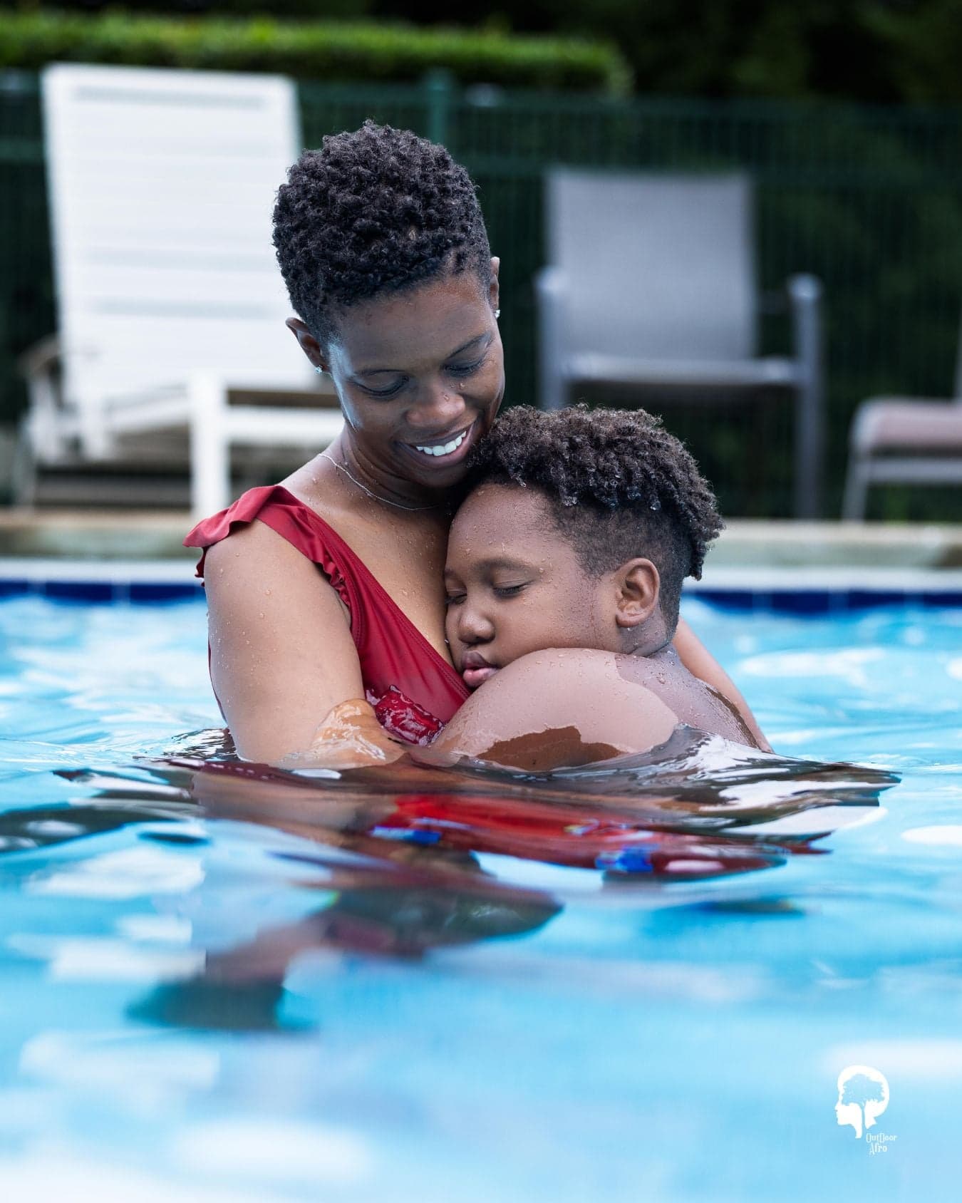 An adult in a red swimsuit stands in a swimming pool, gently hugging a child who rests their head on the adult’s shoulder, both partially submerged in the water
