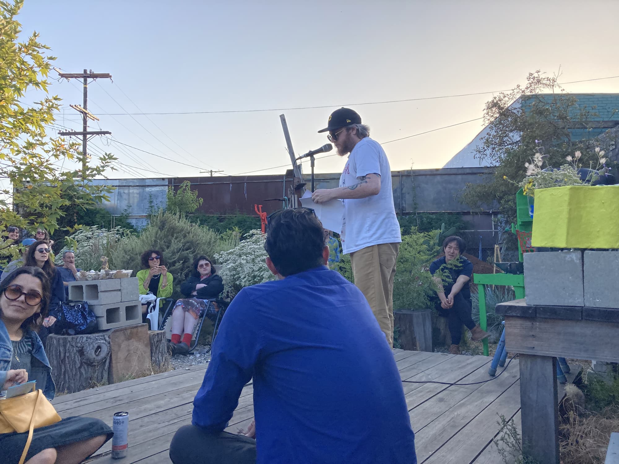 Poet Ed Steck reads from loose leaves of paper onstage to a small crowd at an urban garden at dusk. He wears a ballcap, sunglasses, a grey tee.