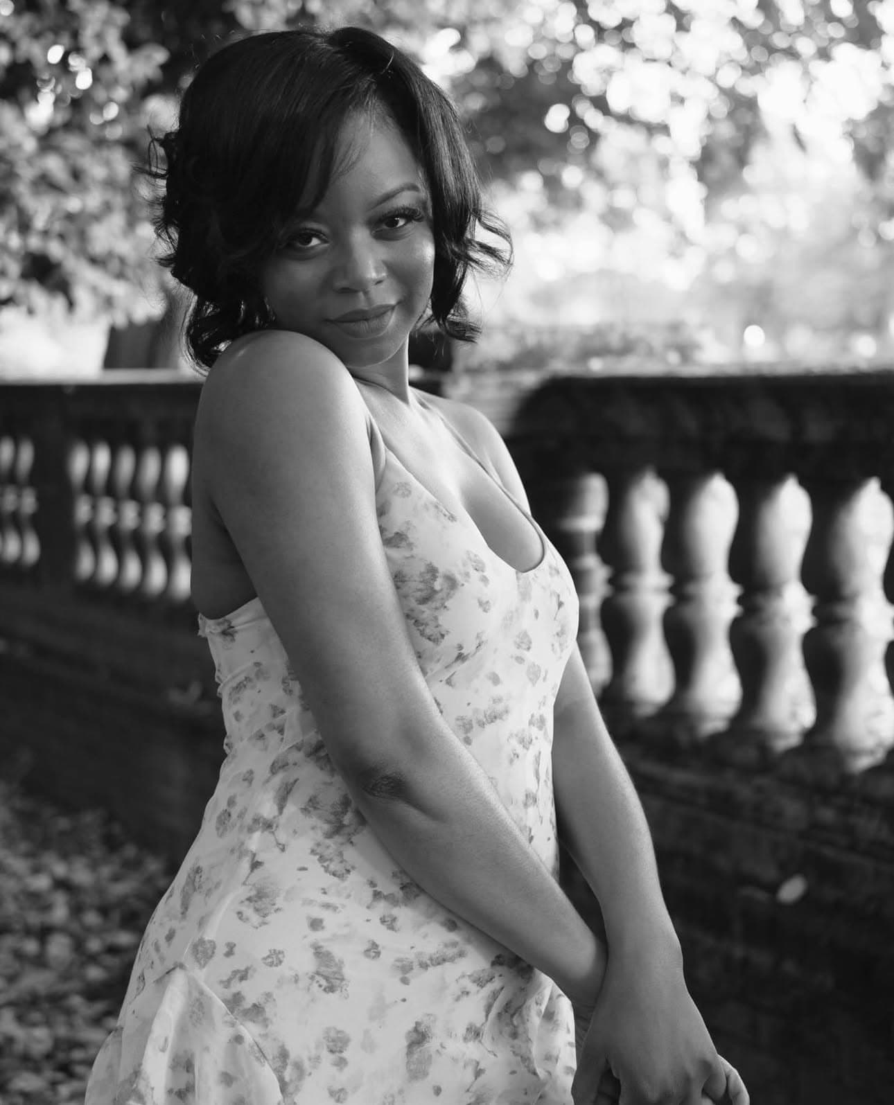 A black-and-white portrait of a Versie Jean, an African American woman with short, wavy hair in a flower-print dress. Her hands are clasped in front of her.