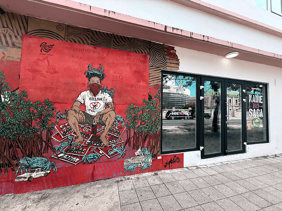 The outside of La Quince, seen from the street. There is a mural on the left side wall by BembaPR with a figure wearing a Abolish Police shirt.